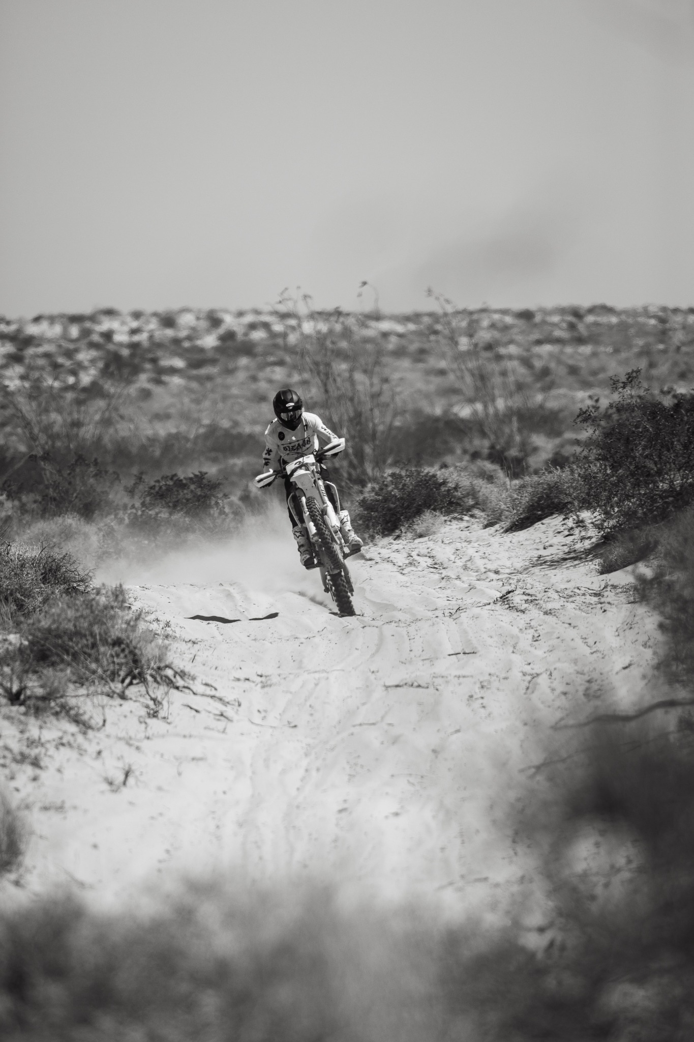 Motorcycle rider approaching along a sandy desert trail.